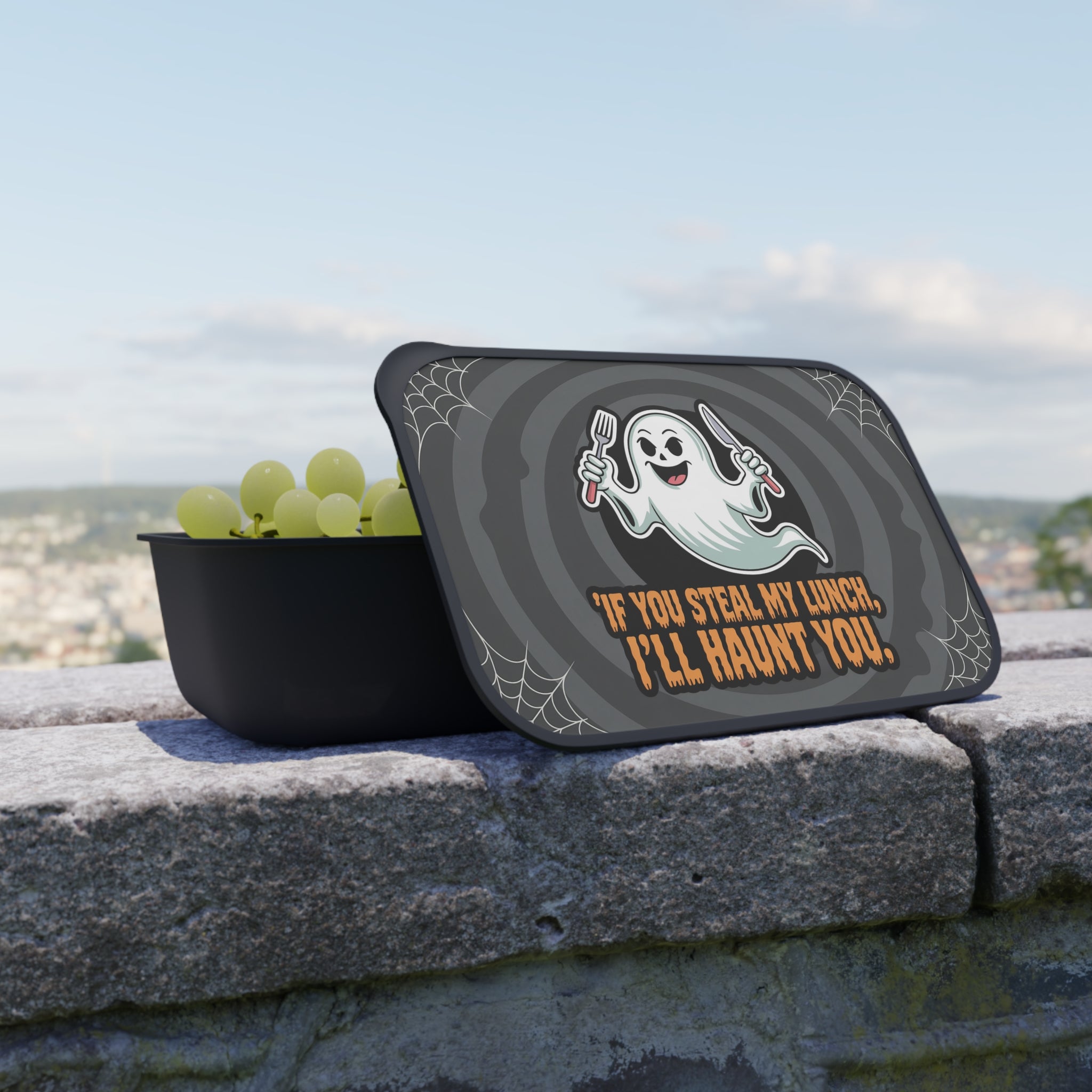 Black lunchbox with lid featuring a ghost and spiderweb design, filled with grapes.