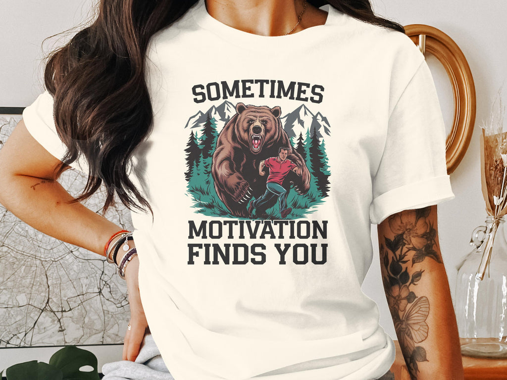 T-shirt graphic of a man running from a bear with text "SOMETIMES MOTIVATION FINDS YOU".