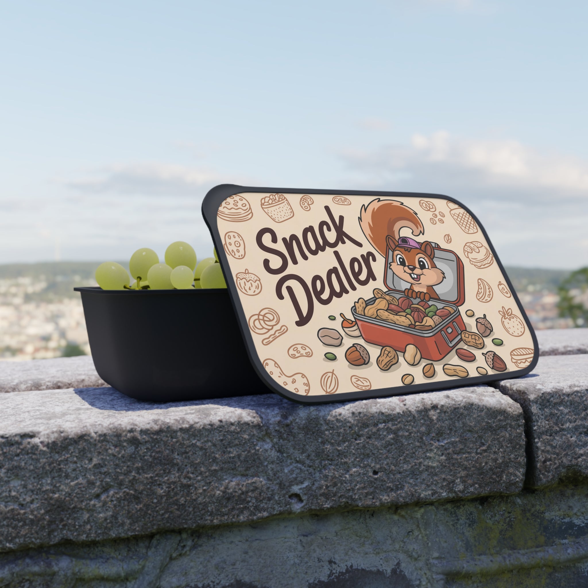 Open black lunchbox with "Snack Dealer" lid filled with grapes and nuts.
