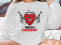Red strawberry with guns, text "THIS IS A STROBERRY" on white sweatshirt.