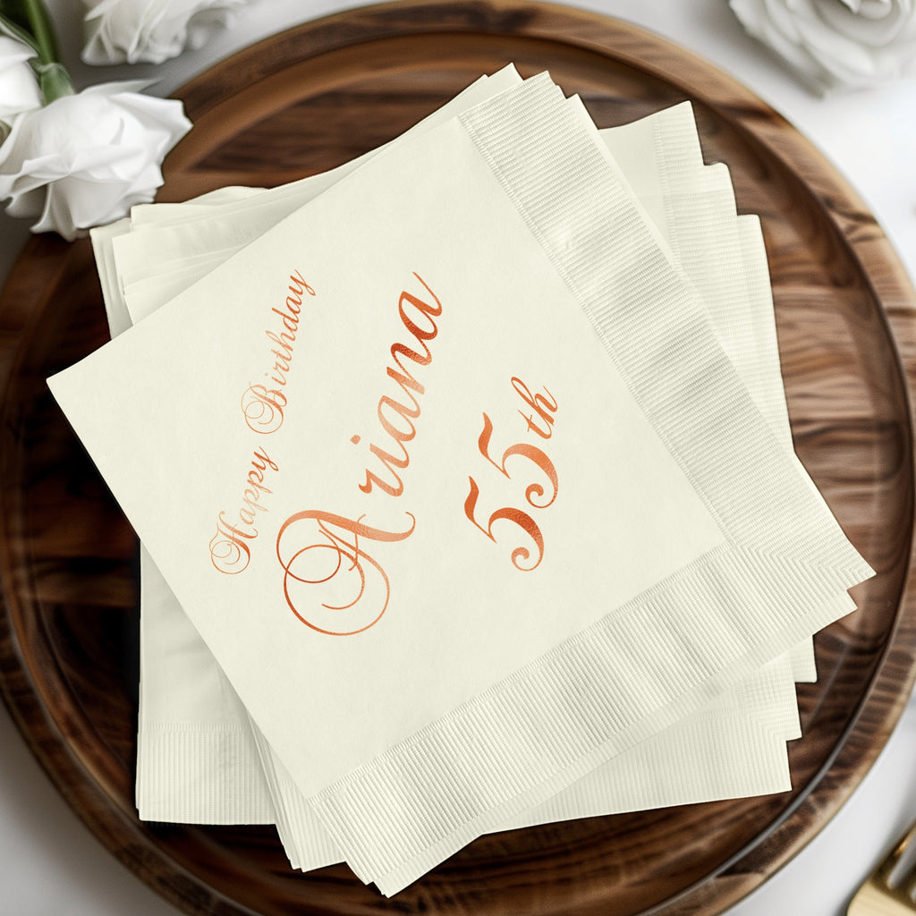 White cocktail napkins with "Happy Birthday Ariana 55th" in rose gold.