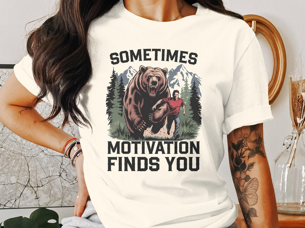 T-shirt graphic: man running from bear in forest, text "SOMETIMES MOTIVATION FINDS YOU".