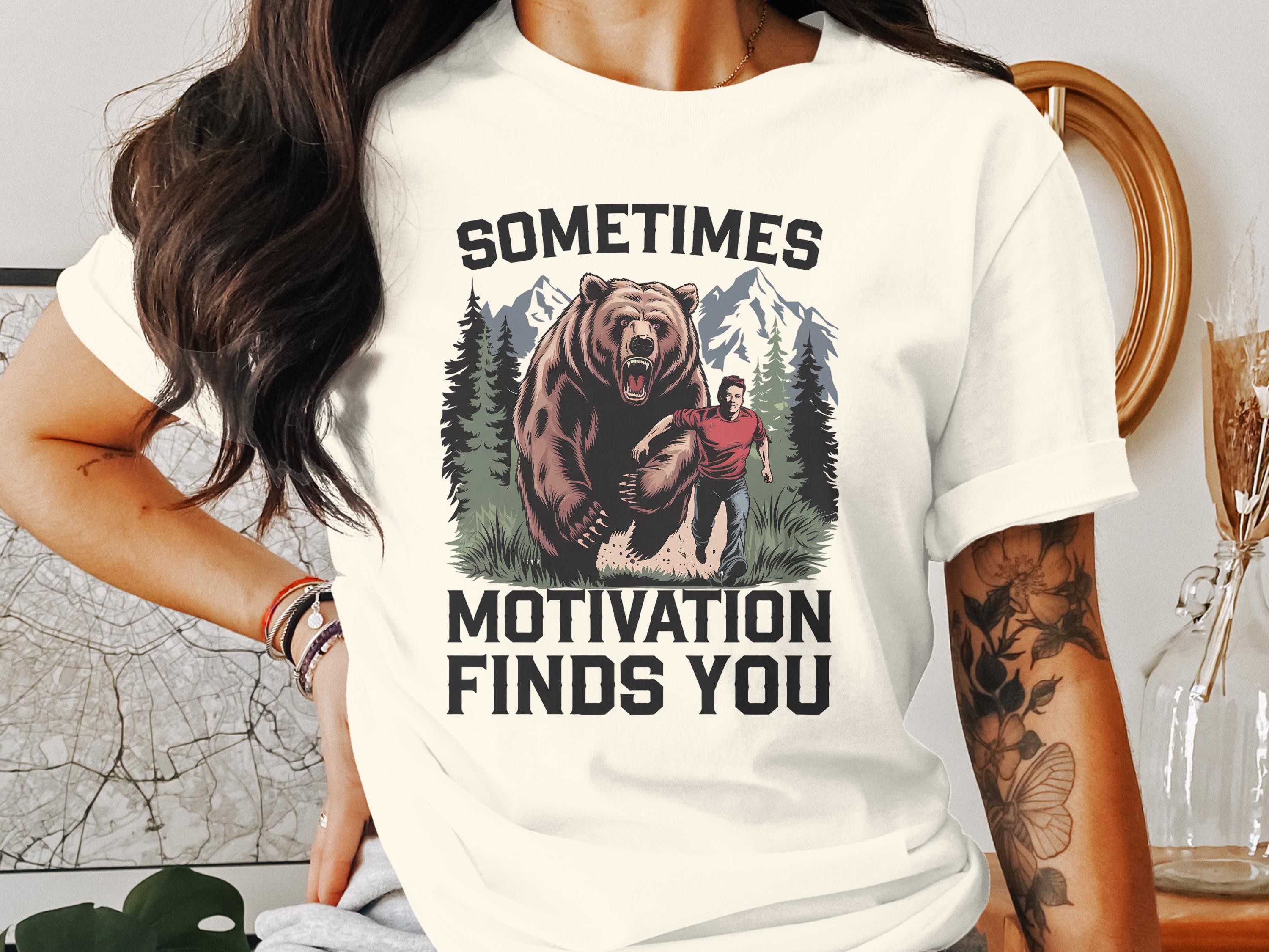 T-shirt graphic: man running from bear in forest, text "SOMETIMES MOTIVATION FINDS YOU".